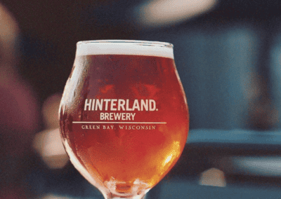 A goblet glass full of amber-colored beer that says Hinterland Brewery, Green Bay, Wisconsin on it sitting outside.