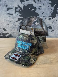 Two camouflage hats, one facing forward with the words "Hinterland Brewery" embroidered on the front, the other facing backwards showing that it is a mesh snapback hat.