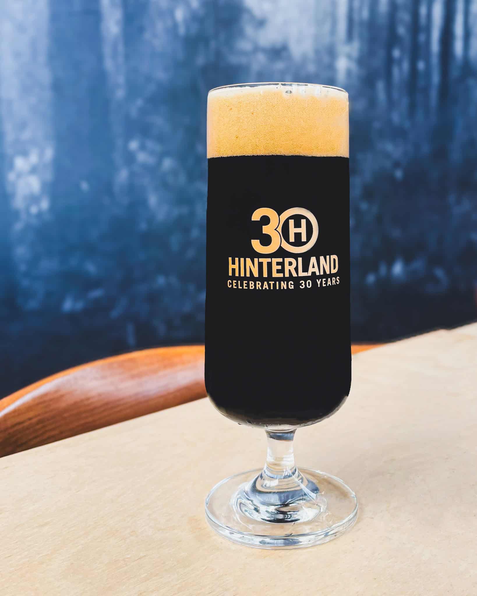 A picture of the Hinterland 30th anniversary glass - a slender, kolsch-looking glass but with a short stem that has the Hinterland 30th anniversary logo in gold ink.