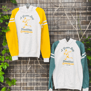 Two hoodies hanging on a fence. One has yellow sleeve and one has green sleeves. They both have a retro football player on them and the phrase "A Green Bay Tradition."