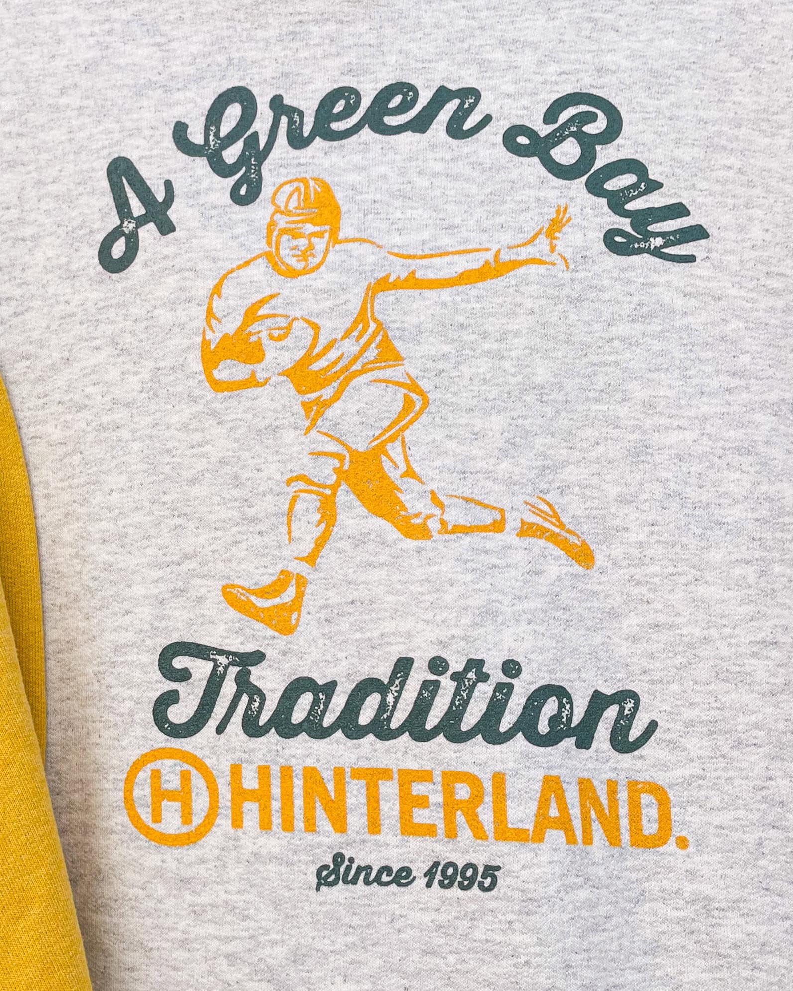 A close up of a graphic on a sweatshirt that has a vintage football player in the center, and has the words "A Green Bay Tradition" around it. It also has the Hinterland Brewery logo.