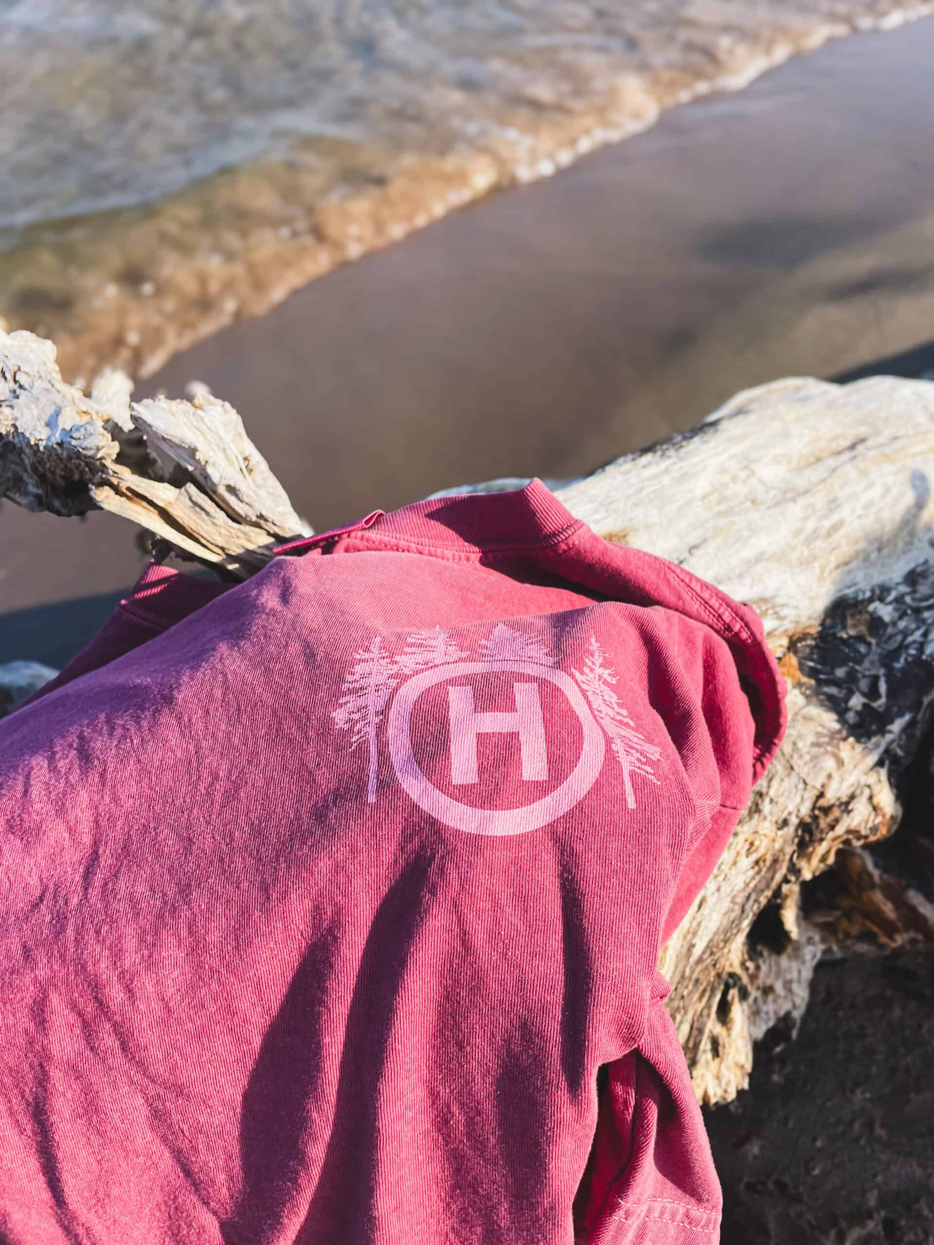A red shirt draped over a piece of drift wood with Lake Michigan in the background. The upper left chest has the Hinterland H logo surrounded by trees.